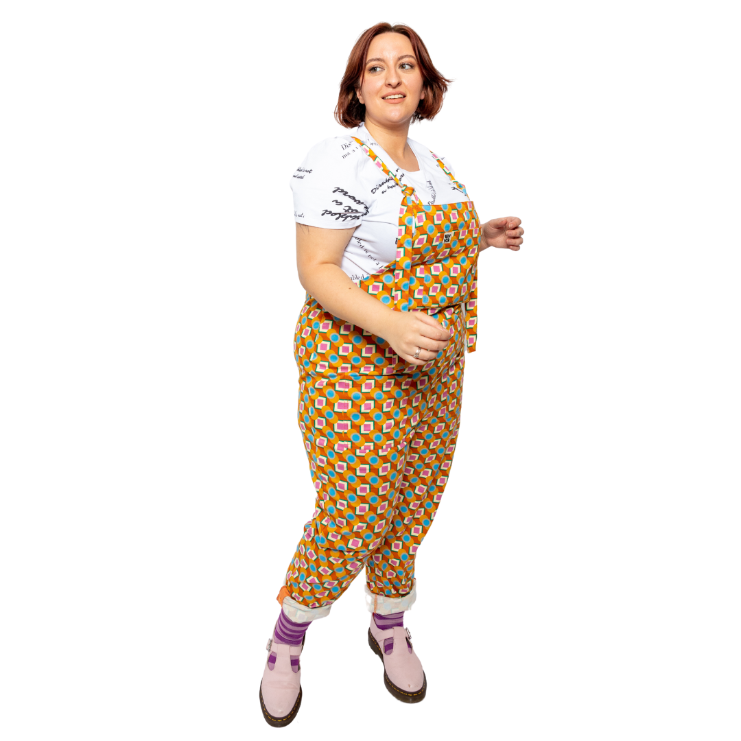 Essie, is wearing a pair of orange and green bold print dungarees with pale pink Dr Martins and purple stripy socks.