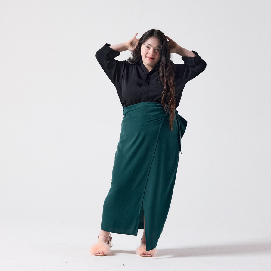 A white woman with long dark brown hair is standing with her hands across her head. She is wearing a forest green wrap skirt in regular fit paired with a black shirt and her own heels.  
