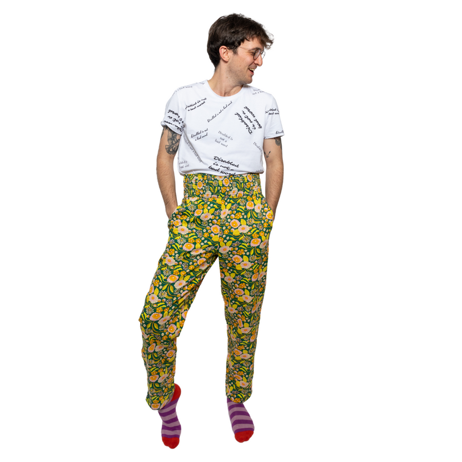 Joshua, a white man with short brown hair, is standing with his weight on one leg and looking to the side. He is wearing green and yellow print trousers with a white t-shirt reading "disabled is not a bad word". He is also wearing stripy purple and pink socks.