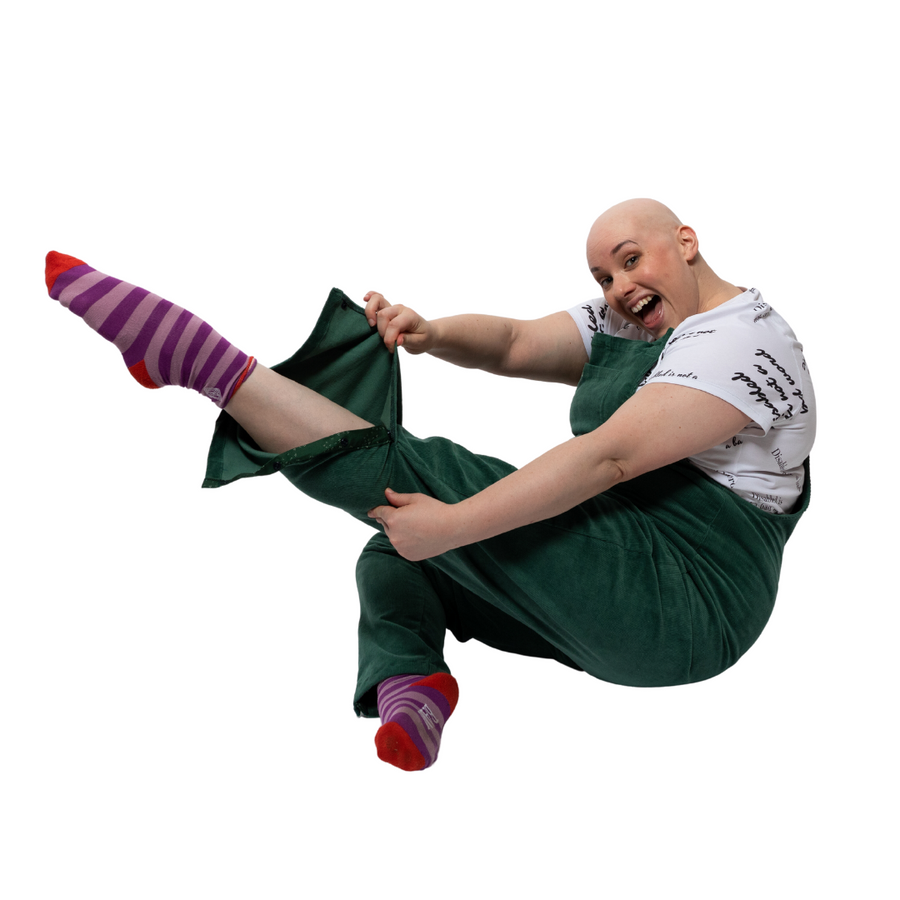 Laura, a white woman with alopecia, is sitting on the floor positioned to the side with one leg pointed out in front of her showing the popper features at the bottom of her trouser leg.  . She is wearing green dungarees with a white t shirt underneath reading "disabled is not a bad word" with stripy purple and pink socks