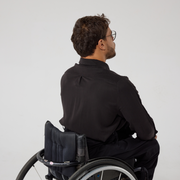 Back view ofMoatez is a male presenting mixed heritage model wearing a black shirt, this is the back view which shows a pleat at the back yoke. He is a manual wheelchair user and has black wire frame glasses on. He's in front of an off white back ground.