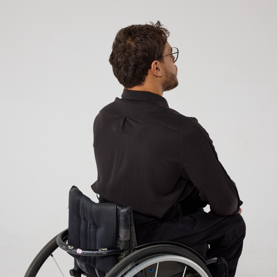 Back view ofMoatez is a male presenting mixed heritage model wearing a black shirt, this is the back view which shows a pleat at the back yoke. He is a manual wheelchair user and has black wire frame glasses on. He's in front of an off white back ground.